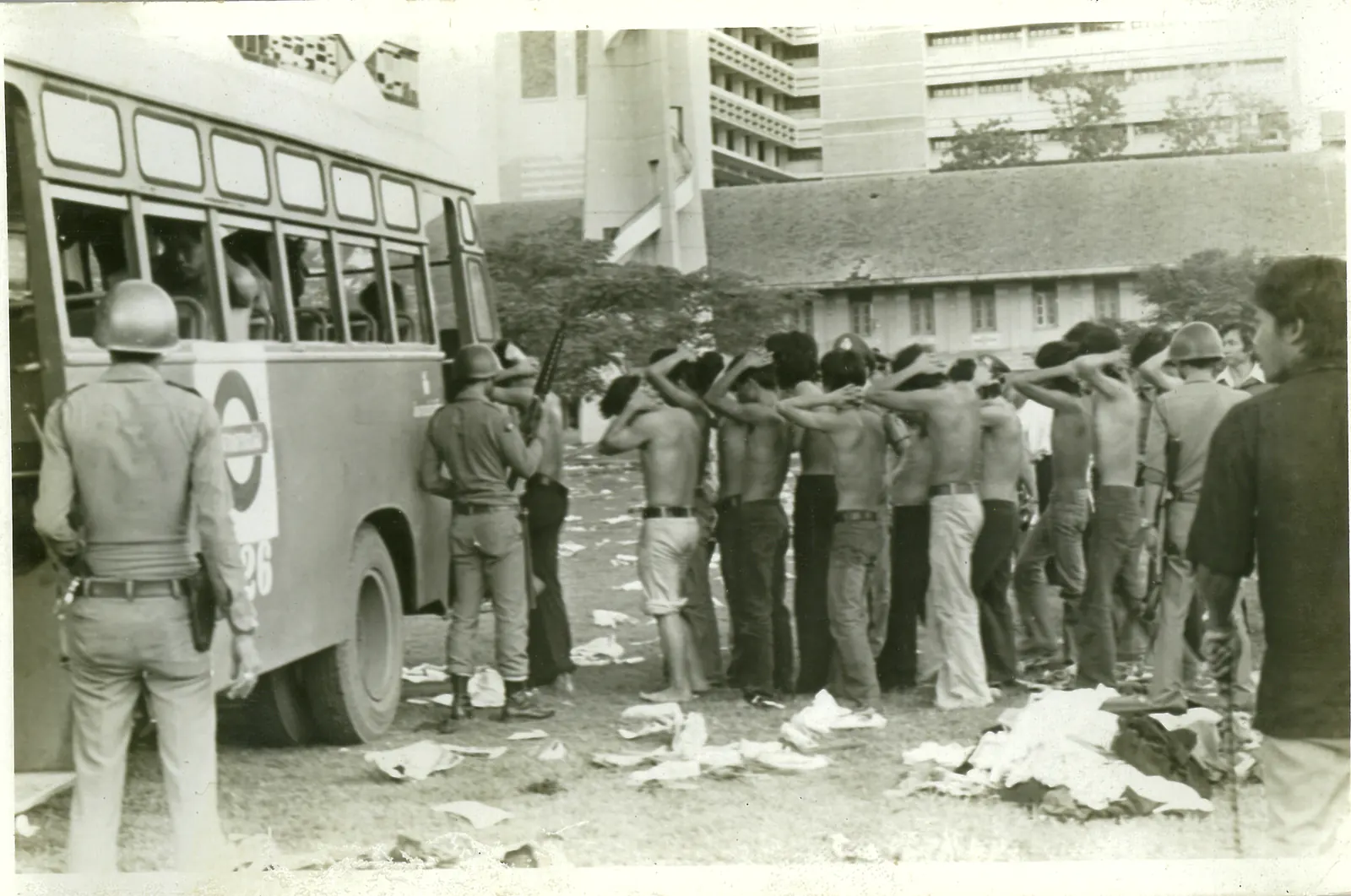 Shirtless detainees stand in a line with their hands on their heads beside a bus, watched by a helmeted policeman.