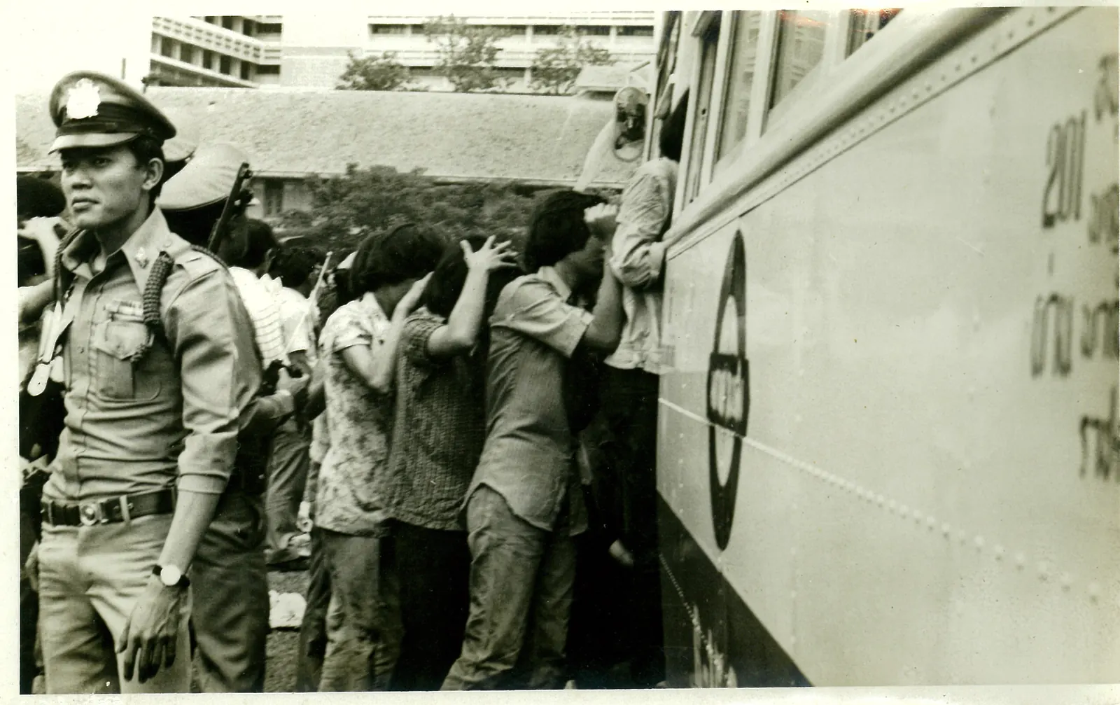 Detainees with their hands on their heads climb onto a bus while a policeman looks on.