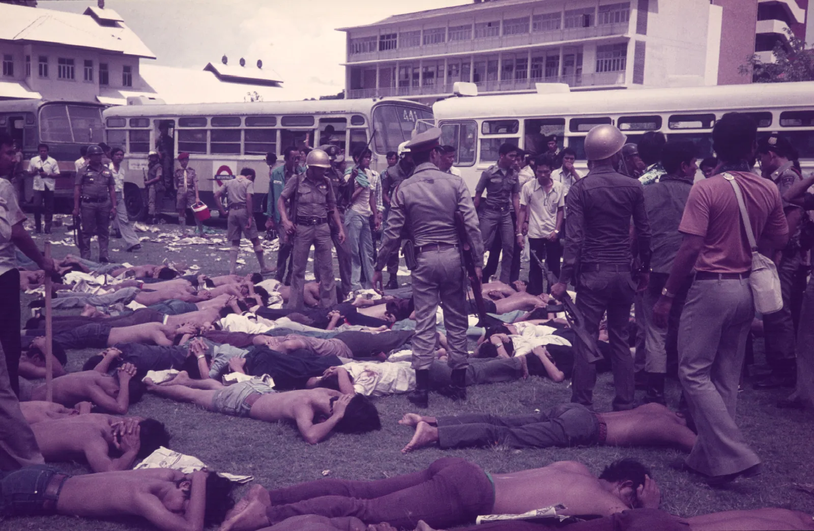 Rows of students lie face down on the grass, many stripped to the waist, while police stand over them in front of parked buses.