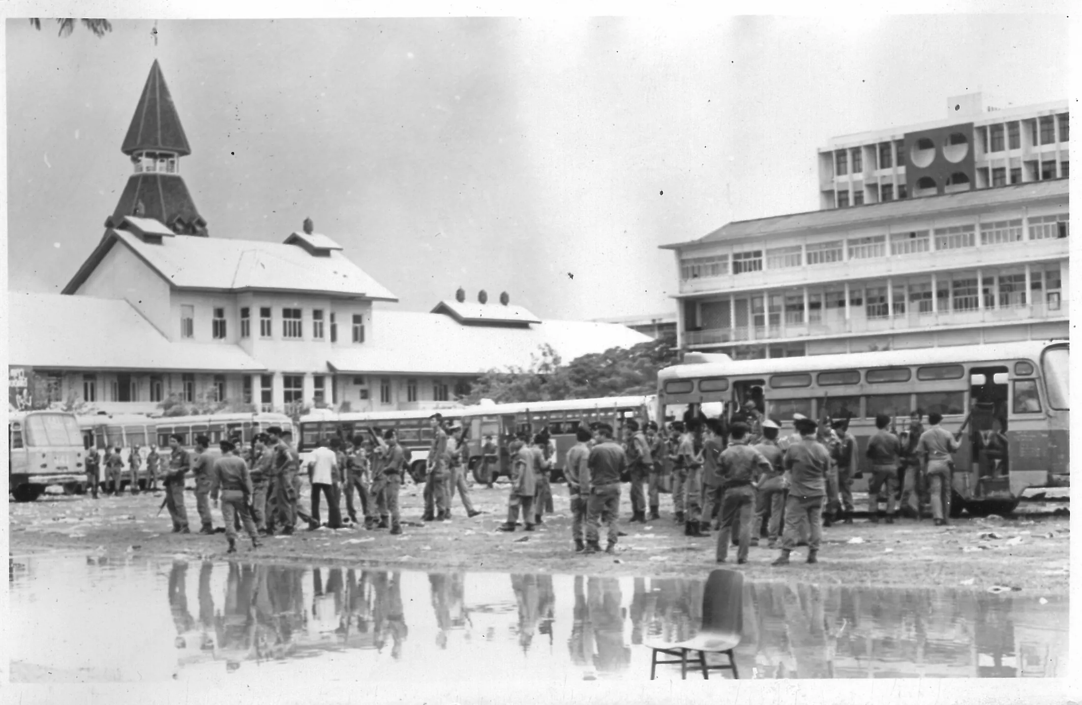 Police with rifles stand among buses on the wet Thammasat football field, with the Dome building and its spire behind them and an empty chair in the foreground.