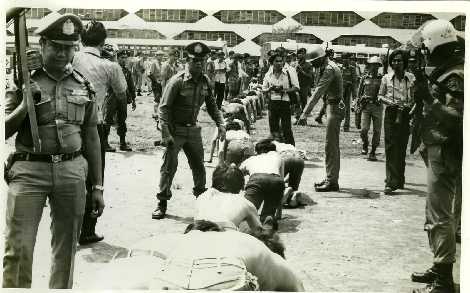 Shirtless young men crawl on hands and knees in a line between police officers holding rifles.