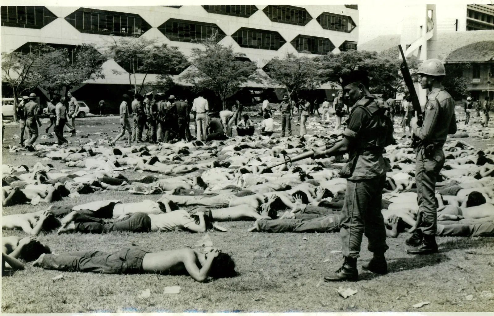 Two armed police stand over rows of students lying face down with their hands behind their heads.