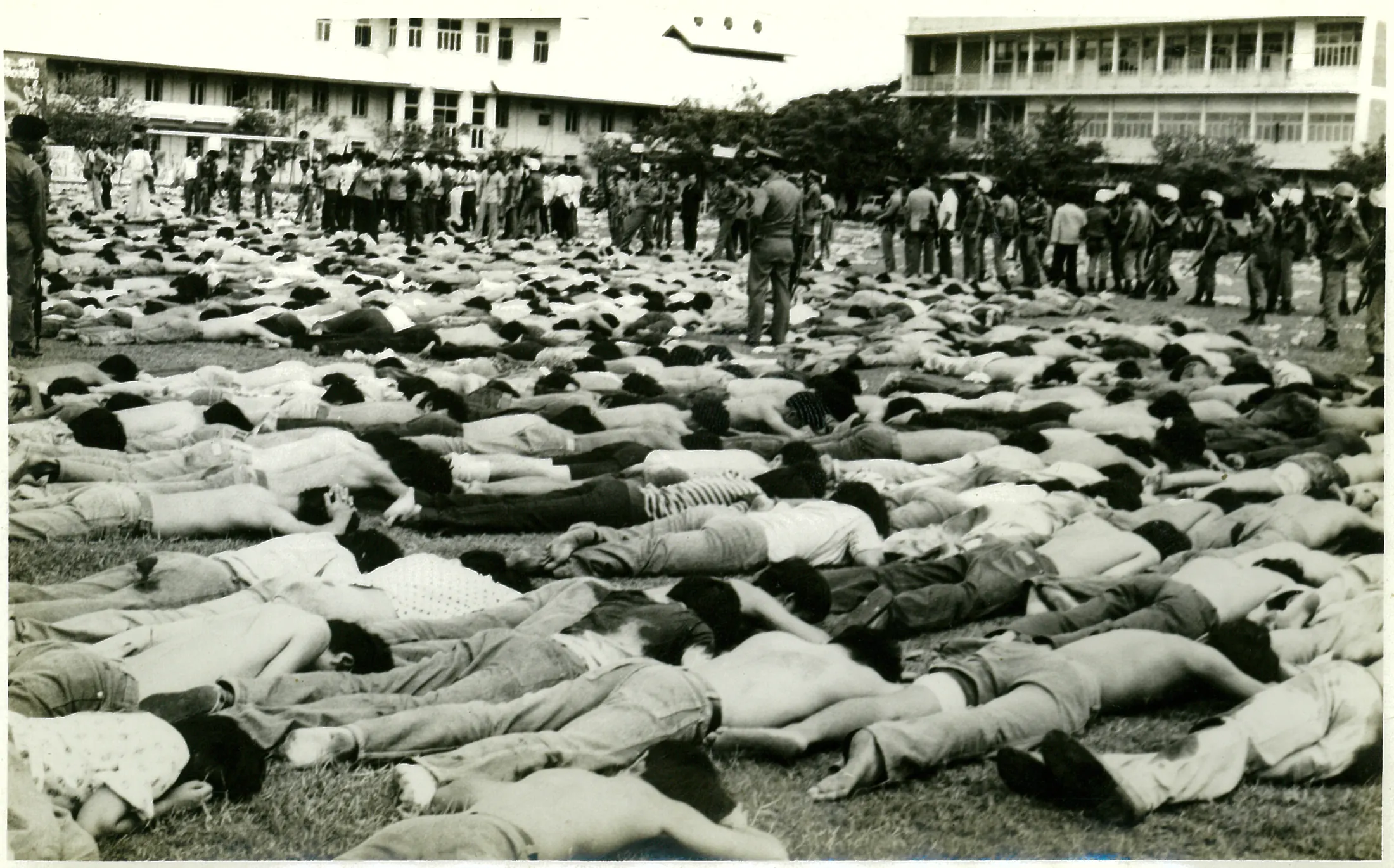 Hundreds of students lie face down in rows across the football field, many stripped to the waist, while police stand along the far side in front of the university buildings.