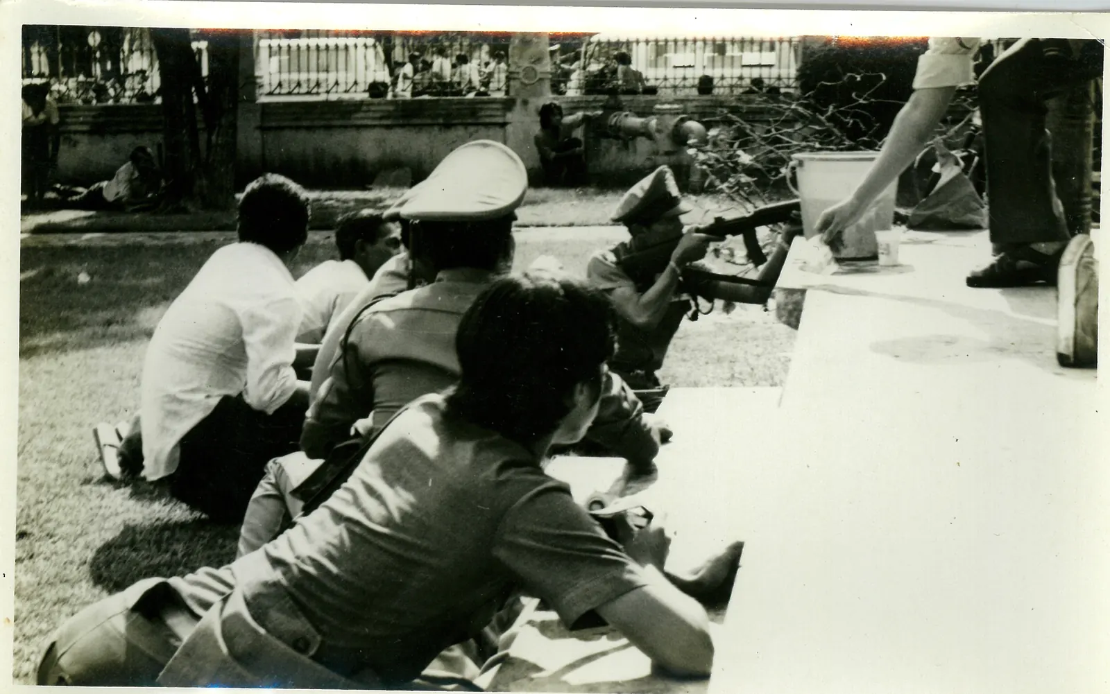 A policeman kneels and aims a rifle while others crouch on the grass beside him.