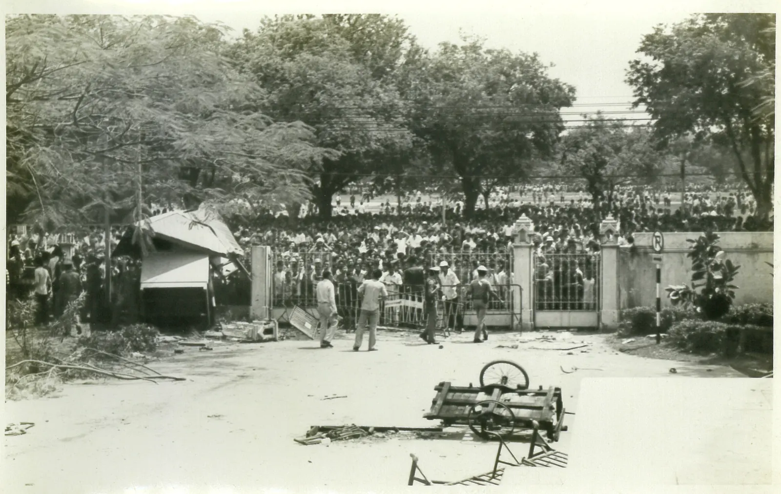 A wrecked sentry box and an overturned cart lie at the main gate, with a dense crowd beyond the railings.