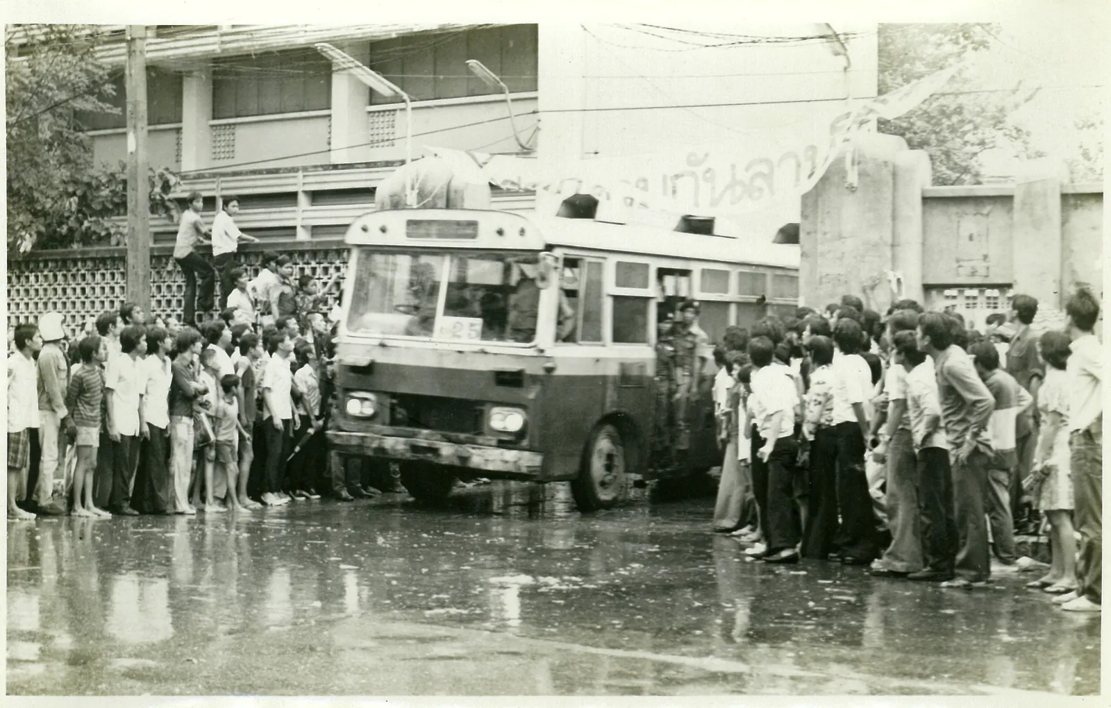 A crowd stands around a city bus pushed against the Tha Prachan gate of Thammasat University.
